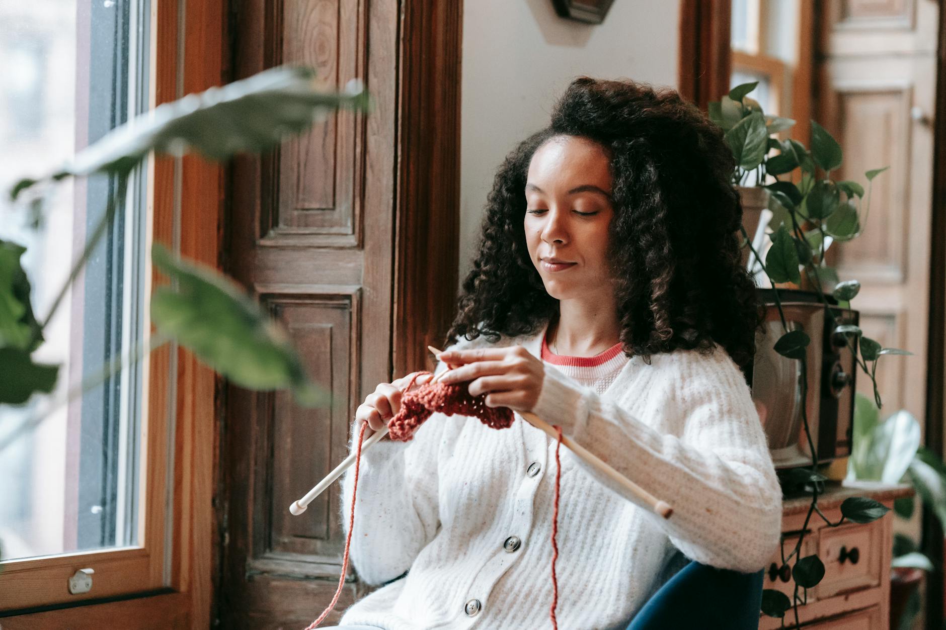 Positive young ethnic female millennial with curly dark hair in casual outfit sitting near window at home and knitting with needles