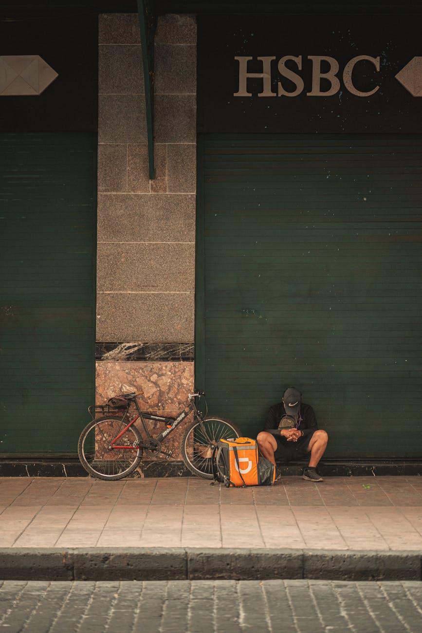 A delivery cyclist rests outside a closed bank with his bike and parcel bag.